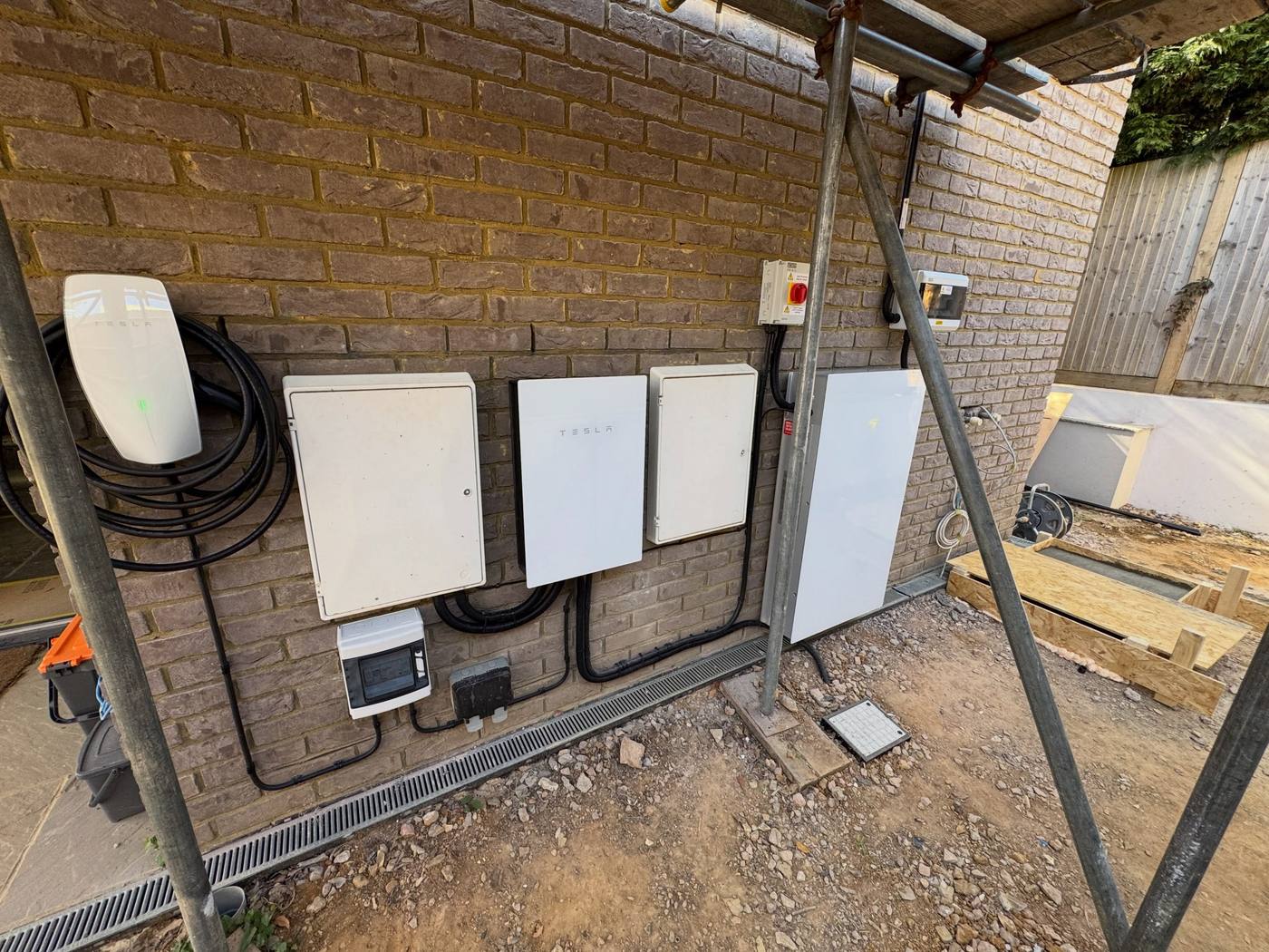 Tesla EV charger alongside Tesla battery and backup equipment and electrical enclosures on an exterior brick wall
